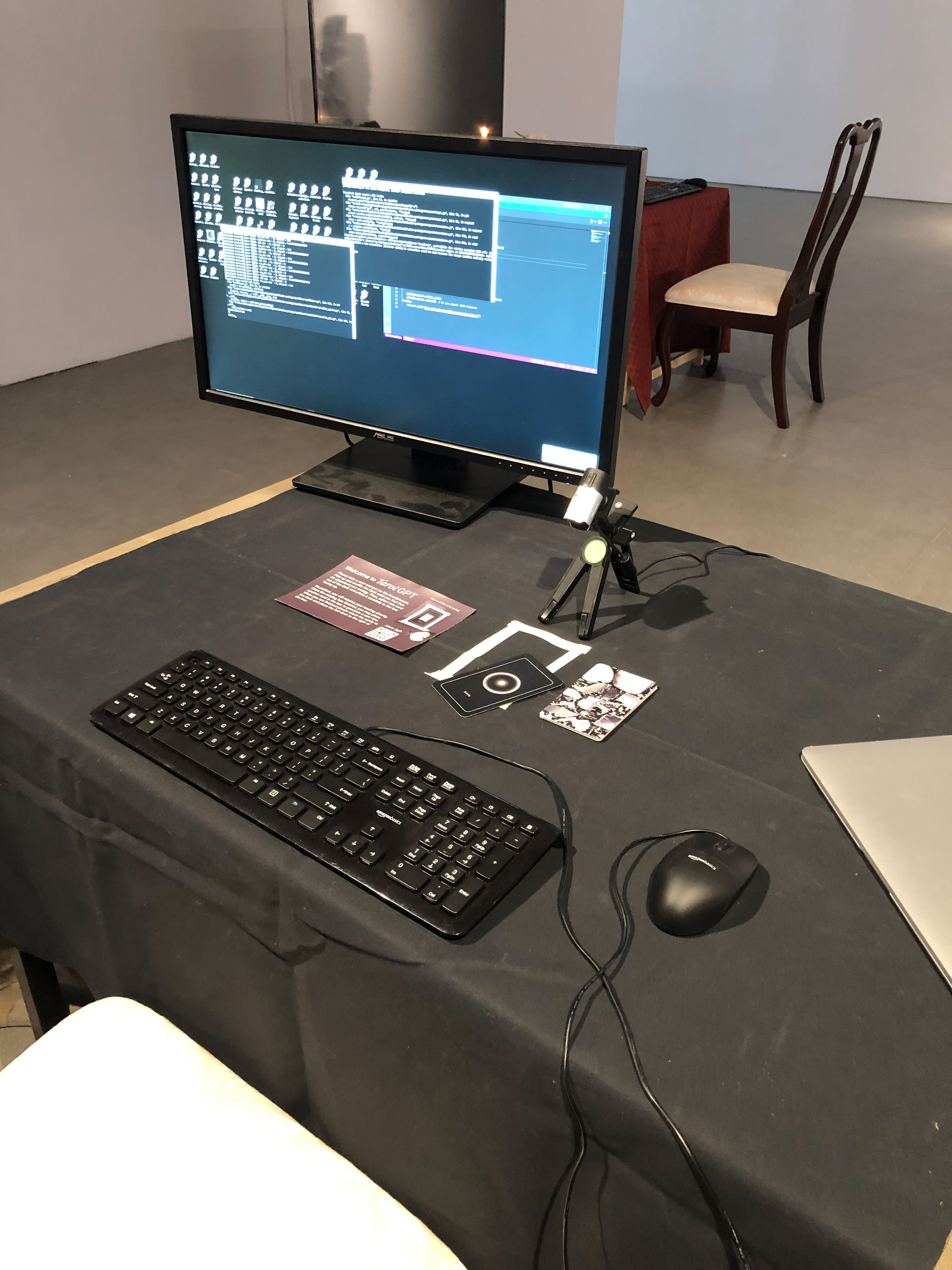 Detailed close up of a table with black table cloth, headphones, a keyboard, a mouse, playing cards and an instruction manual on top. There is a webcam connected to a 6 inch tripod pointing towards the cards.